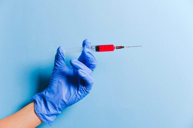 Hand wearing latex glove holds needle in front of a blue background.